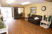 Interior view of a common area in Monte Vista Hills Health Care Center featuring a brown leather couch with a white pillow, two white side tables with lamps and plants, a wall clock, framed artwork, and a wooden chair near a glass door entrance. The floor is wooden and the space is well-lit with ceiling lights.