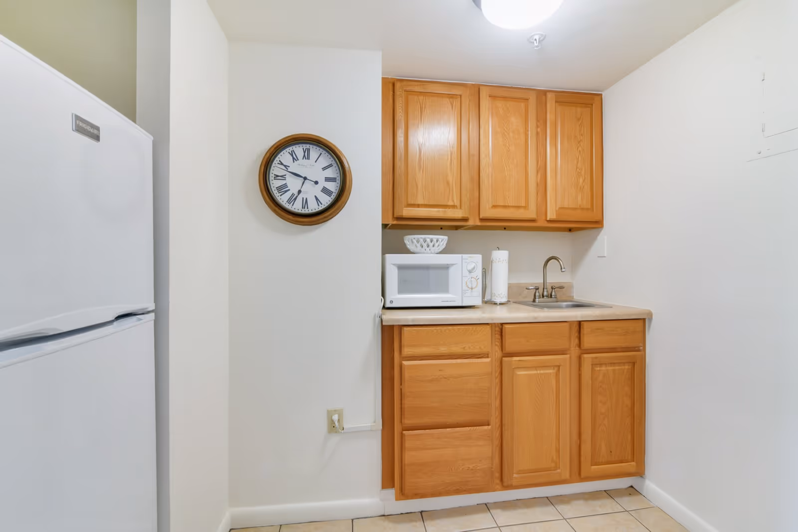 Small kitchenette with wooden cabinets, a microwave and sink on a countertop, a refrigerator to the left, and a round wall clock.