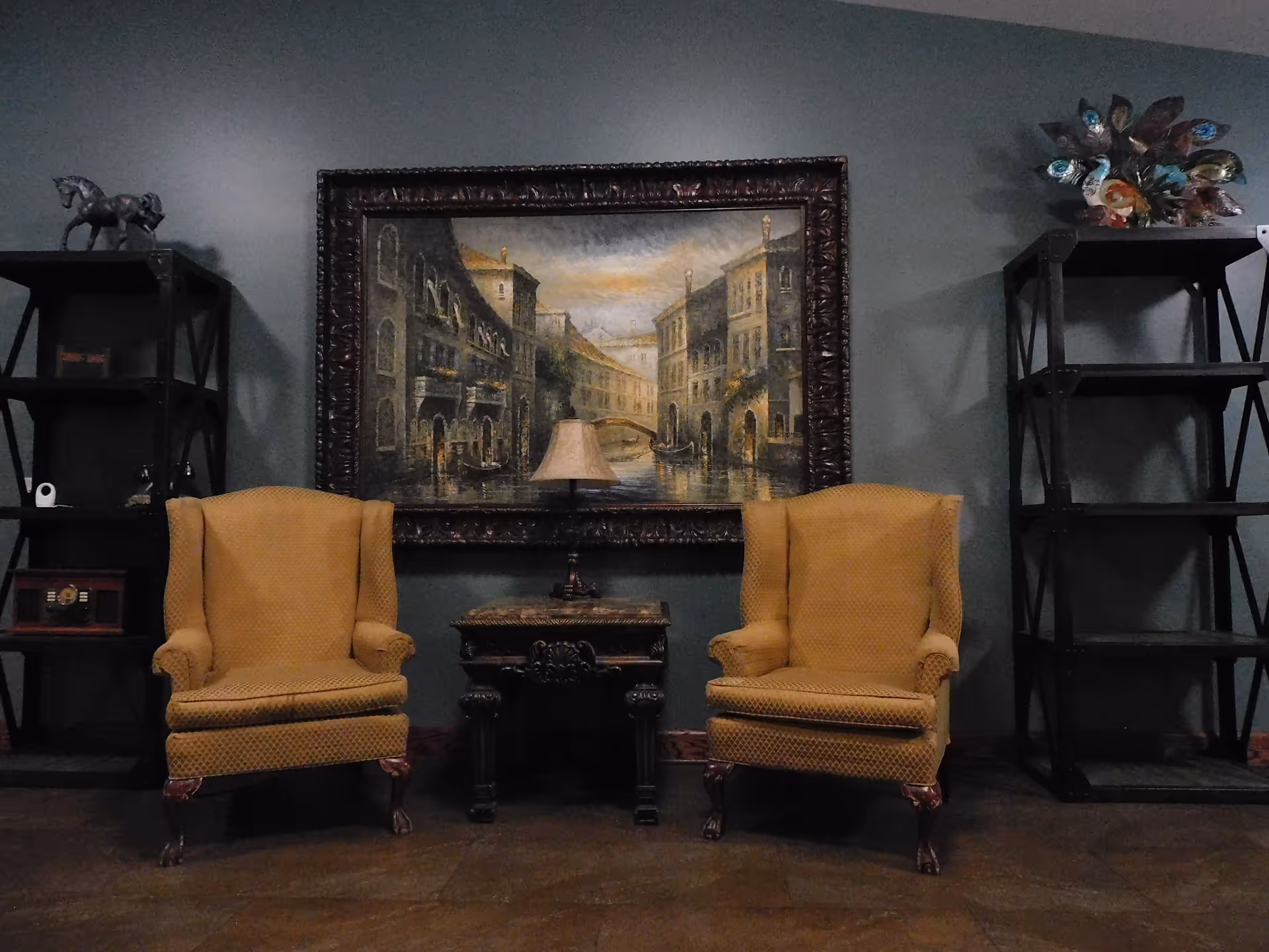 Two yellow upholstered armchairs with wooden legs are positioned on either side of a small ornate wooden table with a lamp on it. Behind the table is a large framed painting depicting a canal scene with buildings and a bridge. On either side of the chairs are black open shelving units, one with decorative items including a horse figurine and a vintage radio, and the other with a colorful peacock sculpture on the top shelf. The walls are painted a muted green color.