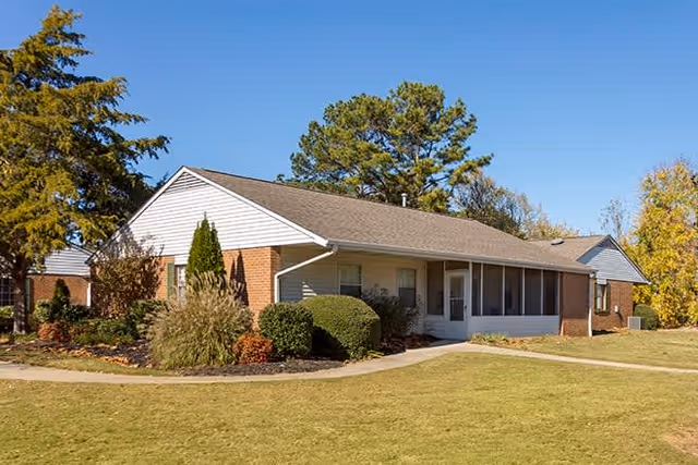Single-story brick building with a gable roof, surrounded by well-maintained landscaping including bushes, trees, and a grassy lawn under a clear blue sky.