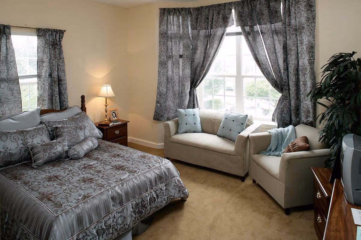 A cozy bedroom with a bed dressed in patterned gray and blue bedding, a wooden nightstand with a lamp and a framed photo, two beige armchairs with blue pillows and a blue throw blanket, large windows with patterned gray curtains, a potted plant, and a wooden dresser with a TV on top.