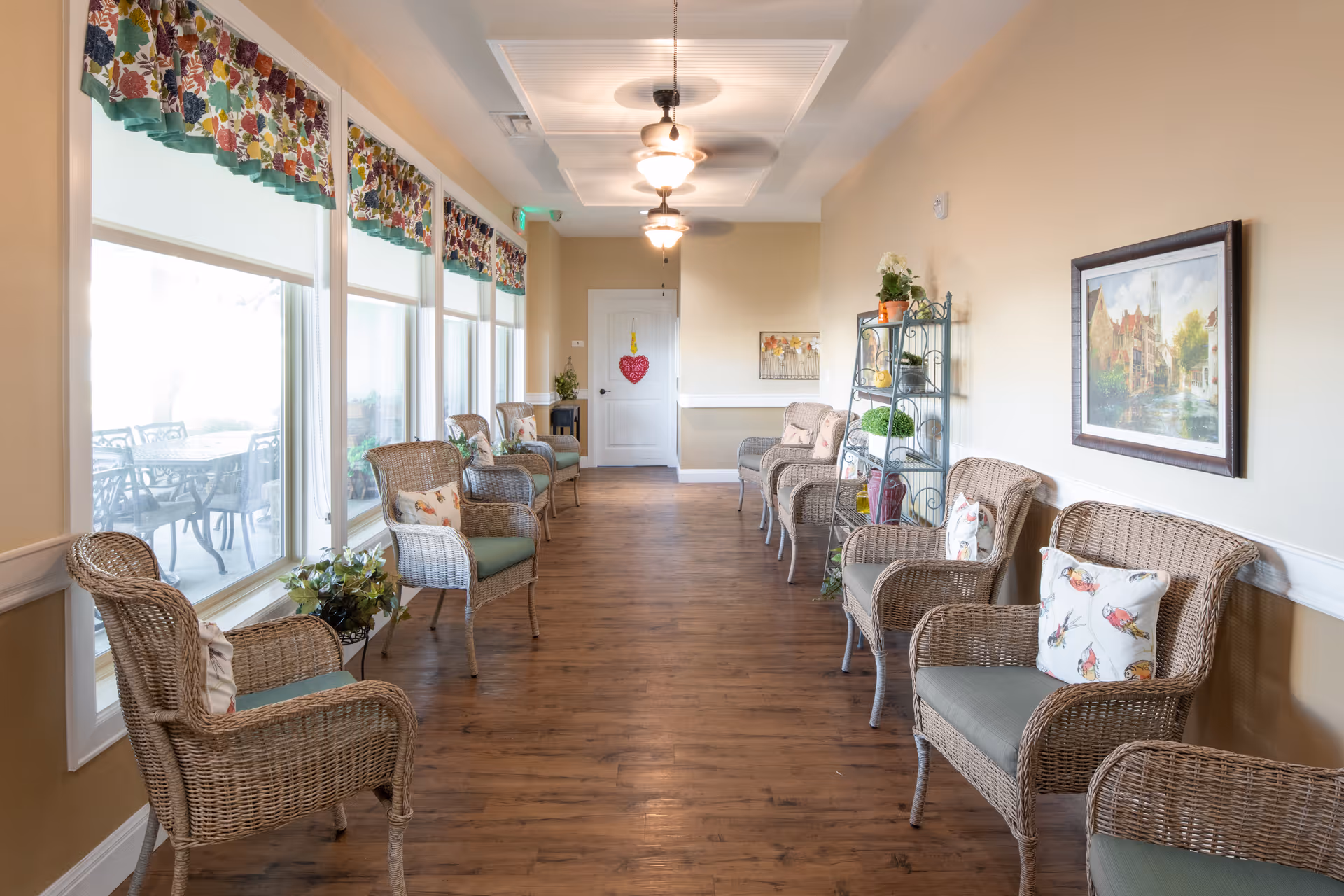 A bright, long hallway with wooden flooring and beige walls, furnished with wicker chairs with cushions and decorative pillows on both sides. Large windows with floral valances line the left side, allowing natural light to fill the space. Ceiling fans with lights hang from the ceiling, and a metal shelving unit with plants and decorative items is on the right wall. A framed painting is also visible on the right wall.