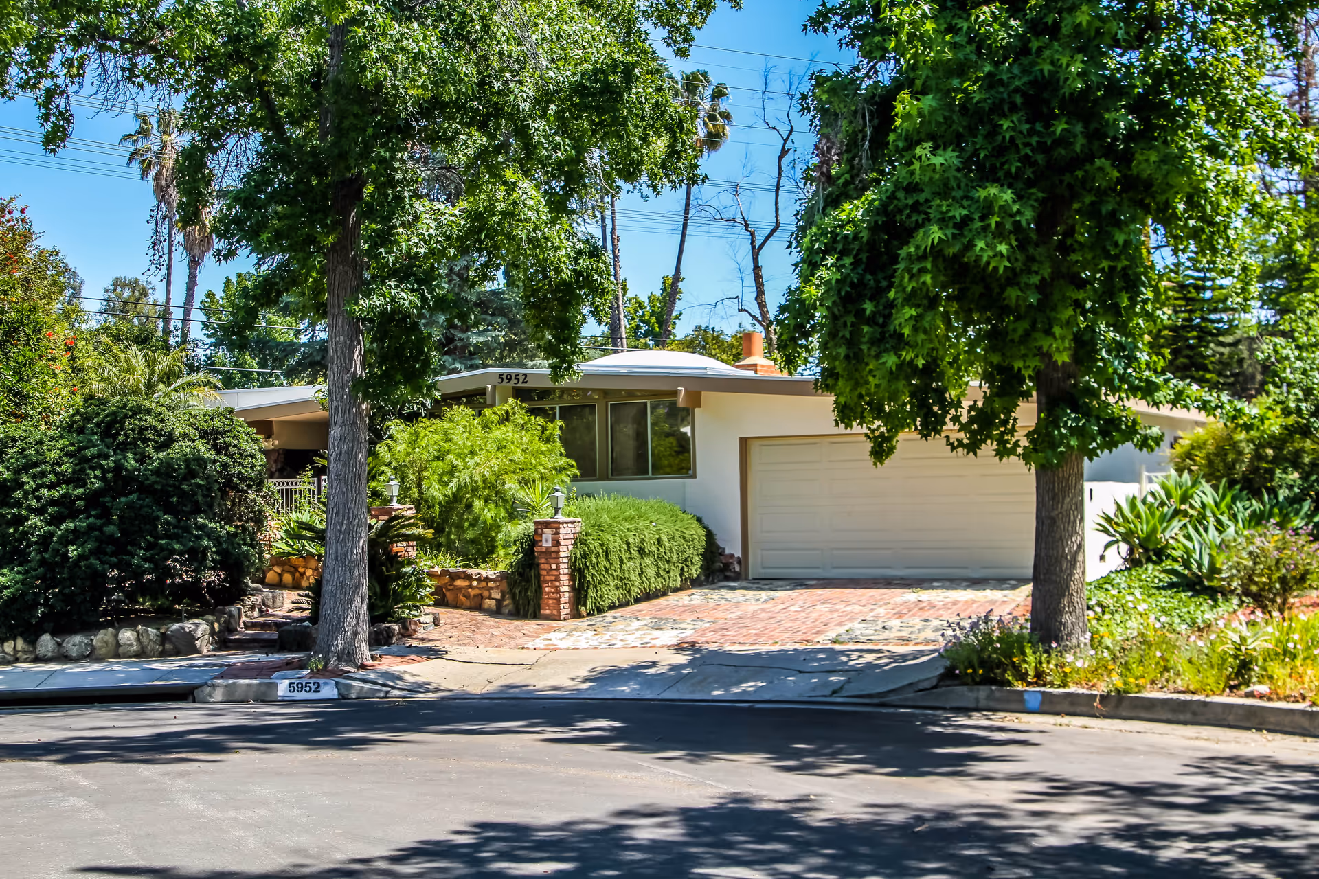 Exterior view of a single-story residential building with a white garage door, surrounded by lush green trees and bushes. The driveway and walkway are made of brick and stone, and the house number 5952 is visible on the curb and above the garage. The sky is clear and blue.