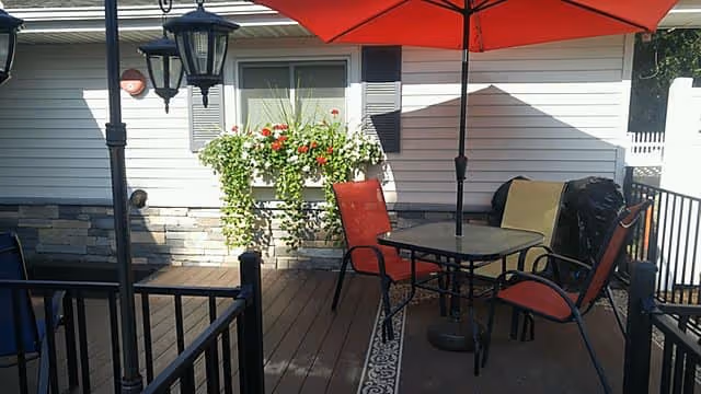Outdoor patio area with a table and four chairs, two of which have red cushions. A large red umbrella provides shade. There is a window with flower boxes filled with green plants and red flowers on the wall of the building. A black grill covered with a beige cloth is visible in the background. The patio has wooden flooring and black railing.