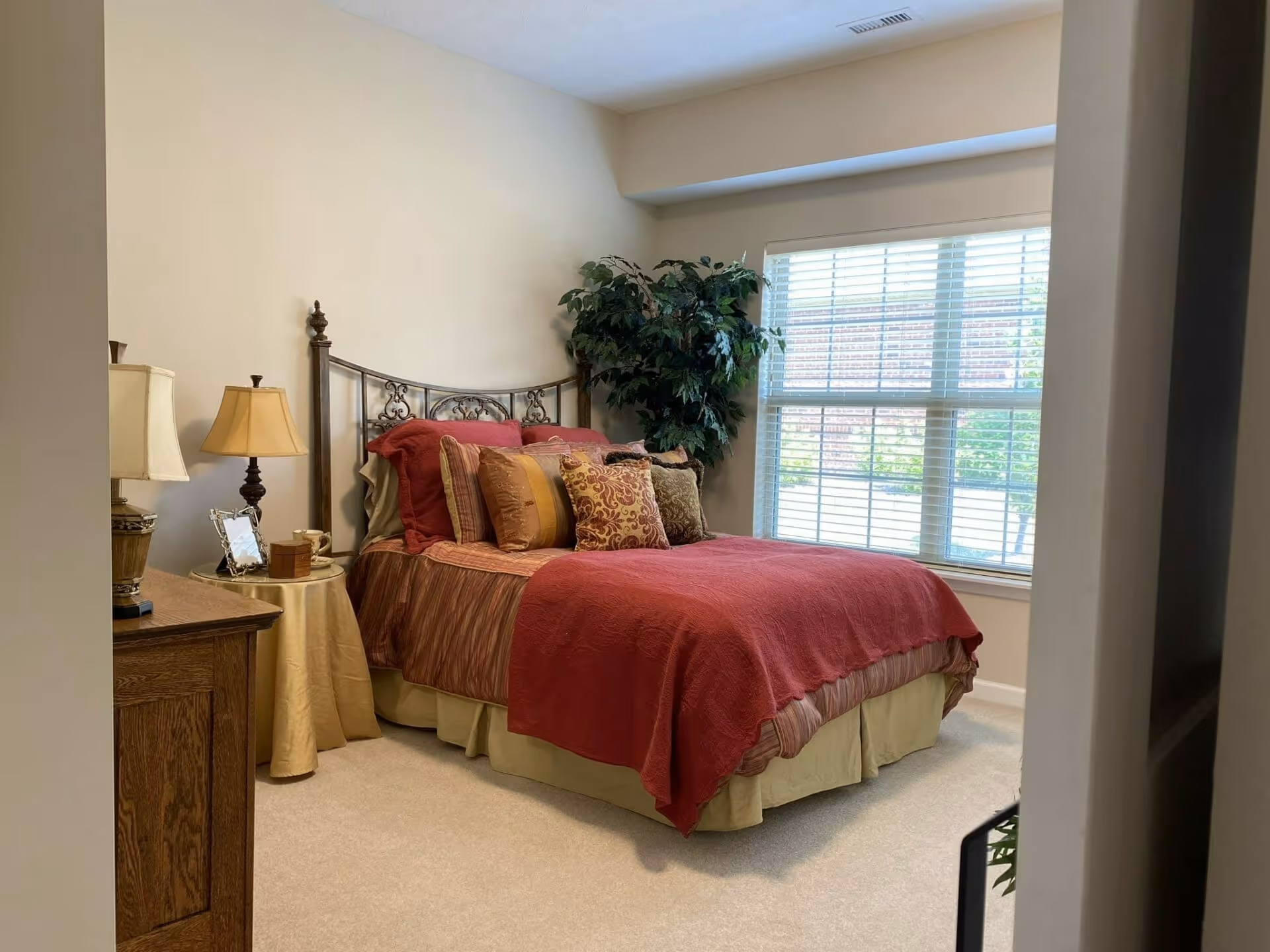 A cozy bedroom with a neatly made bed featuring red and patterned pillows and a red bedspread. There is a wooden dresser with a lamp and picture frame on the left side, and a large window with white blinds letting in natural light. A green potted plant is placed near the window.