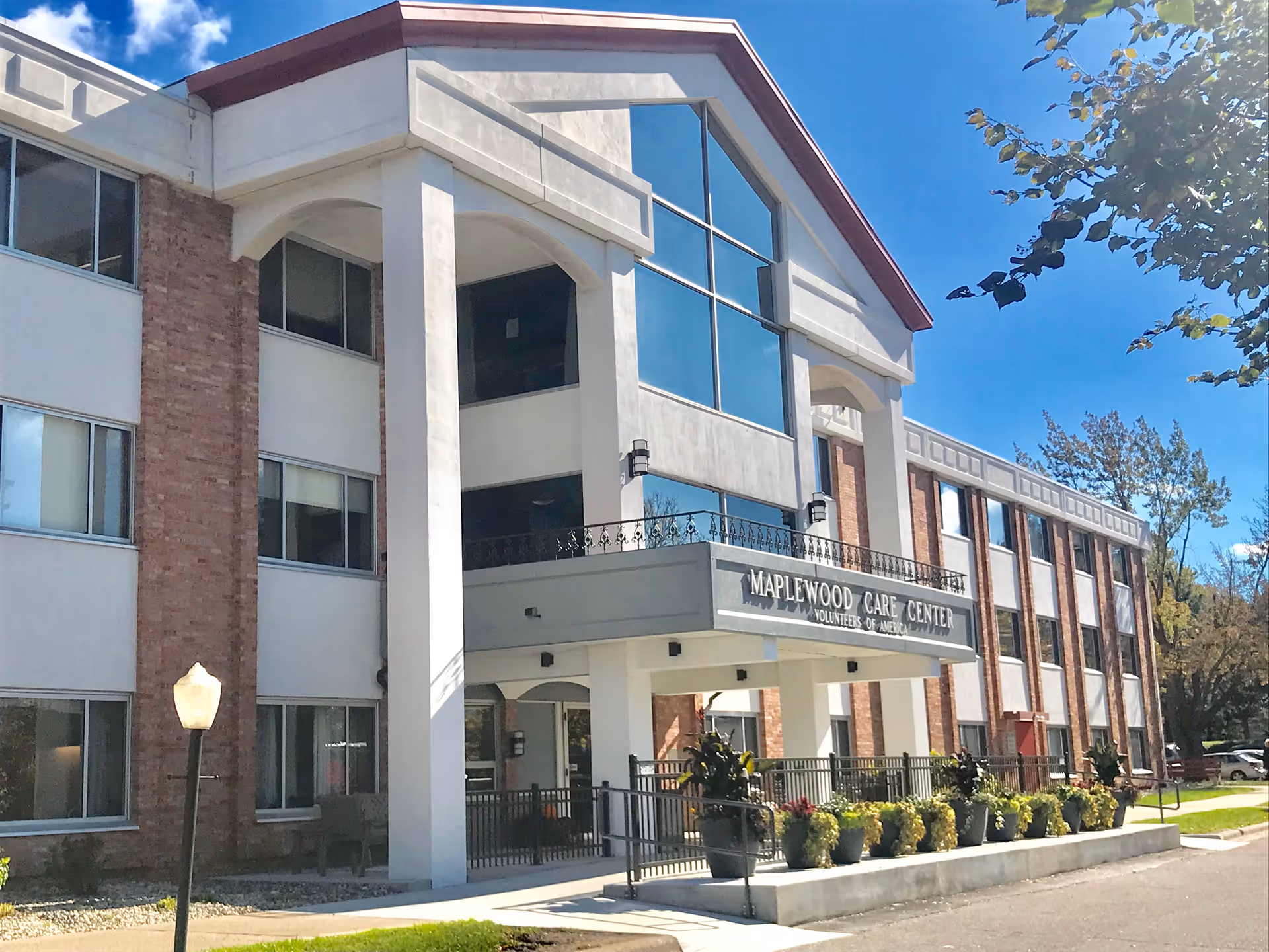 Exterior view of Maplewood Care Center, a multi-story building with large windows and a covered entrance. There are potted plants along the front walkway and a street lamp on the left side. The sky is clear and blue with some trees in the background.