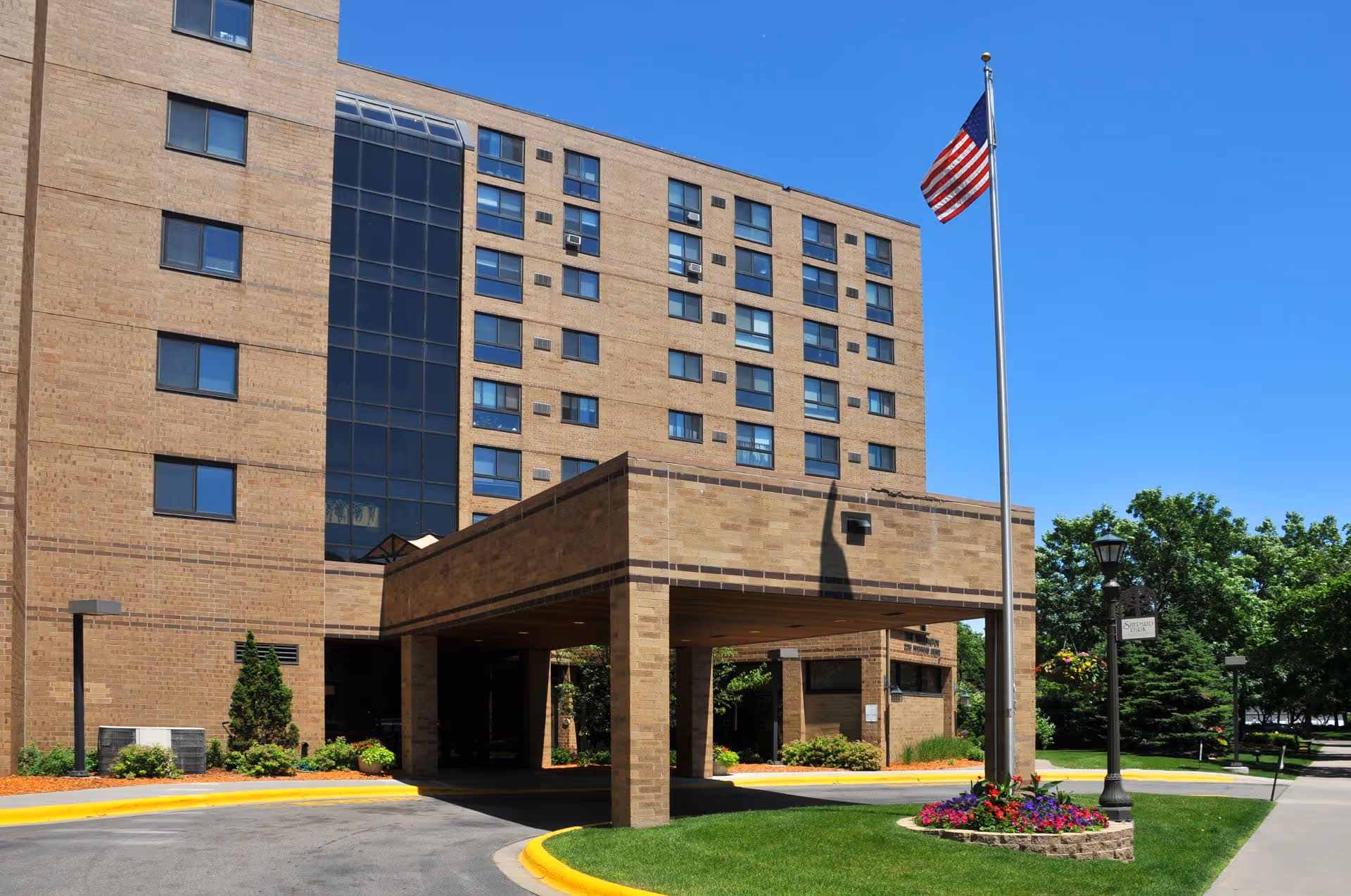 Exterior view of a multi-story senior living facility building with a covered entrance, an American flag on a flagpole, and a landscaped area with flowers and trees under a clear blue sky.