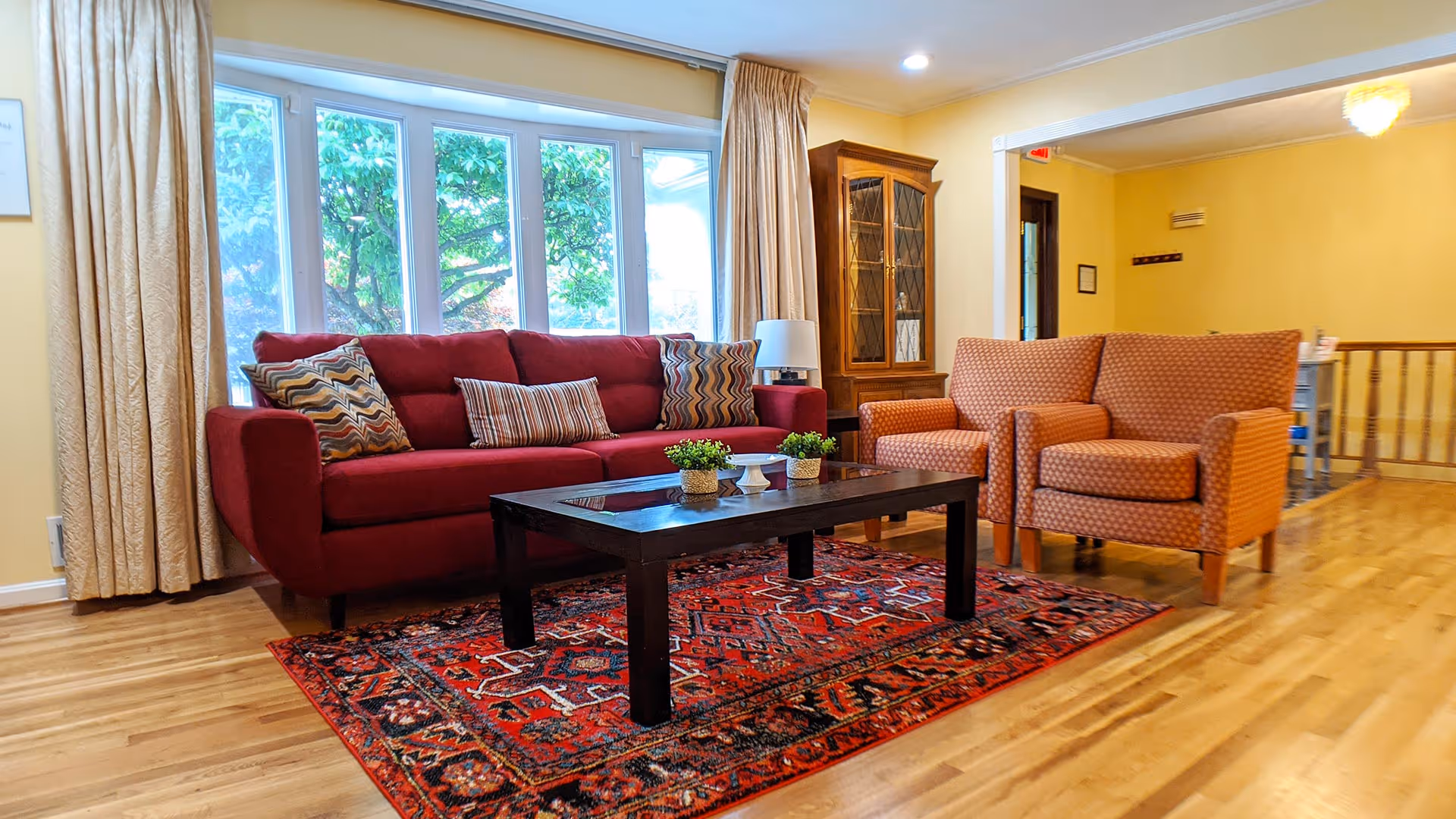 A cozy living room with a large window showing green trees outside. The room features a red sofa with patterned cushions, two orange armchairs, a dark wooden coffee table with small potted plants, a red patterned rug, light wooden flooring, beige curtains, and a wooden cabinet with glass doors.