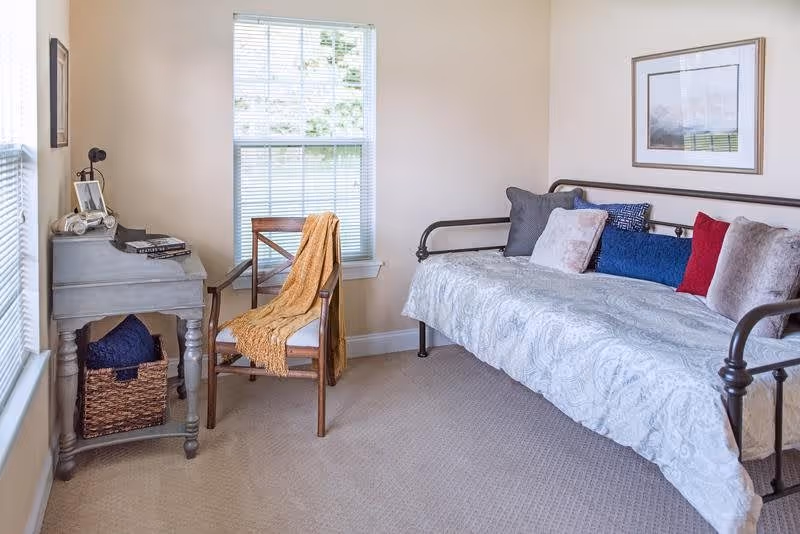 A cozy room with a daybed covered in a light patterned bedspread and several colorful pillows. Next to the bed is a wooden chair draped with a mustard yellow throw blanket. A small gray desk with books and a lamp sits by the window, and a woven basket is placed underneath the desk. The room has beige walls and carpeted floor, with a framed picture hanging above the daybed.