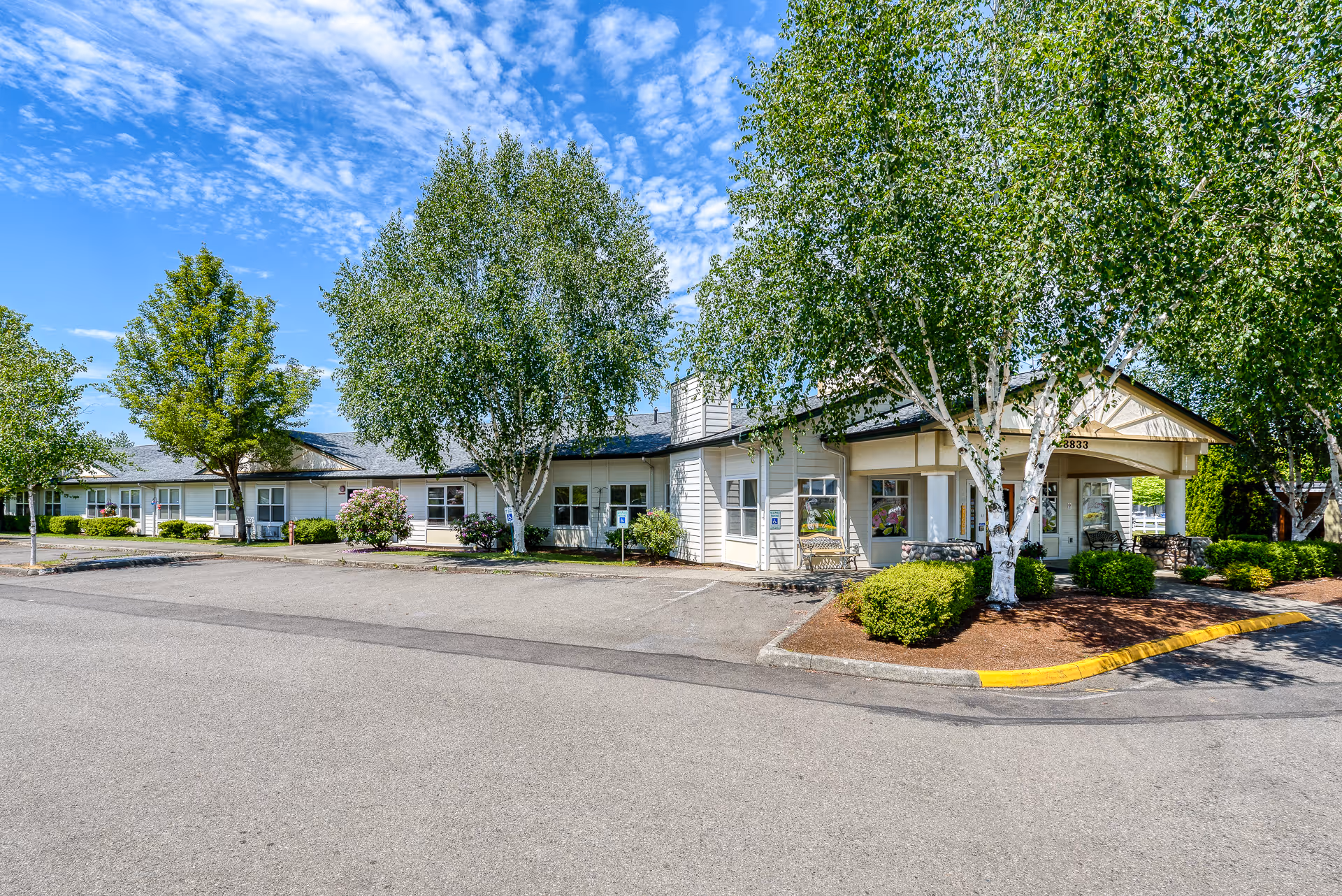 Exterior view of Heritage House Buckley Assisted Living & Memory Care building with a parking lot in front, surrounded by trees and bushes under a partly cloudy blue sky.