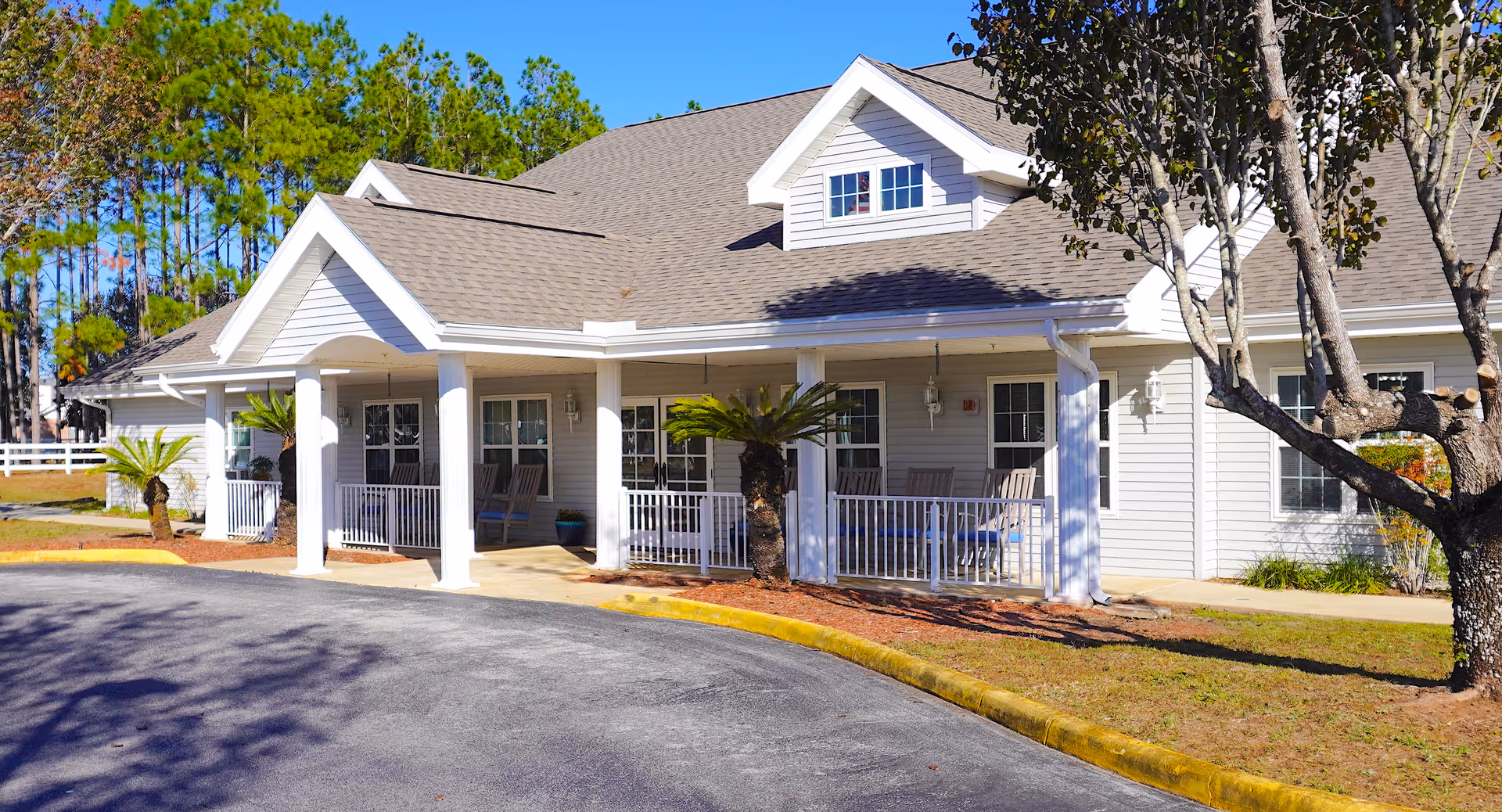 Exterior view of a single-story building with a covered porch supported by white columns. The building has light gray siding, multiple windows, and a gabled roof. There are several rocking chairs on the porch and small palm trees planted near the entrance. The scene is set on a sunny day with a clear blue sky and trees in the background.