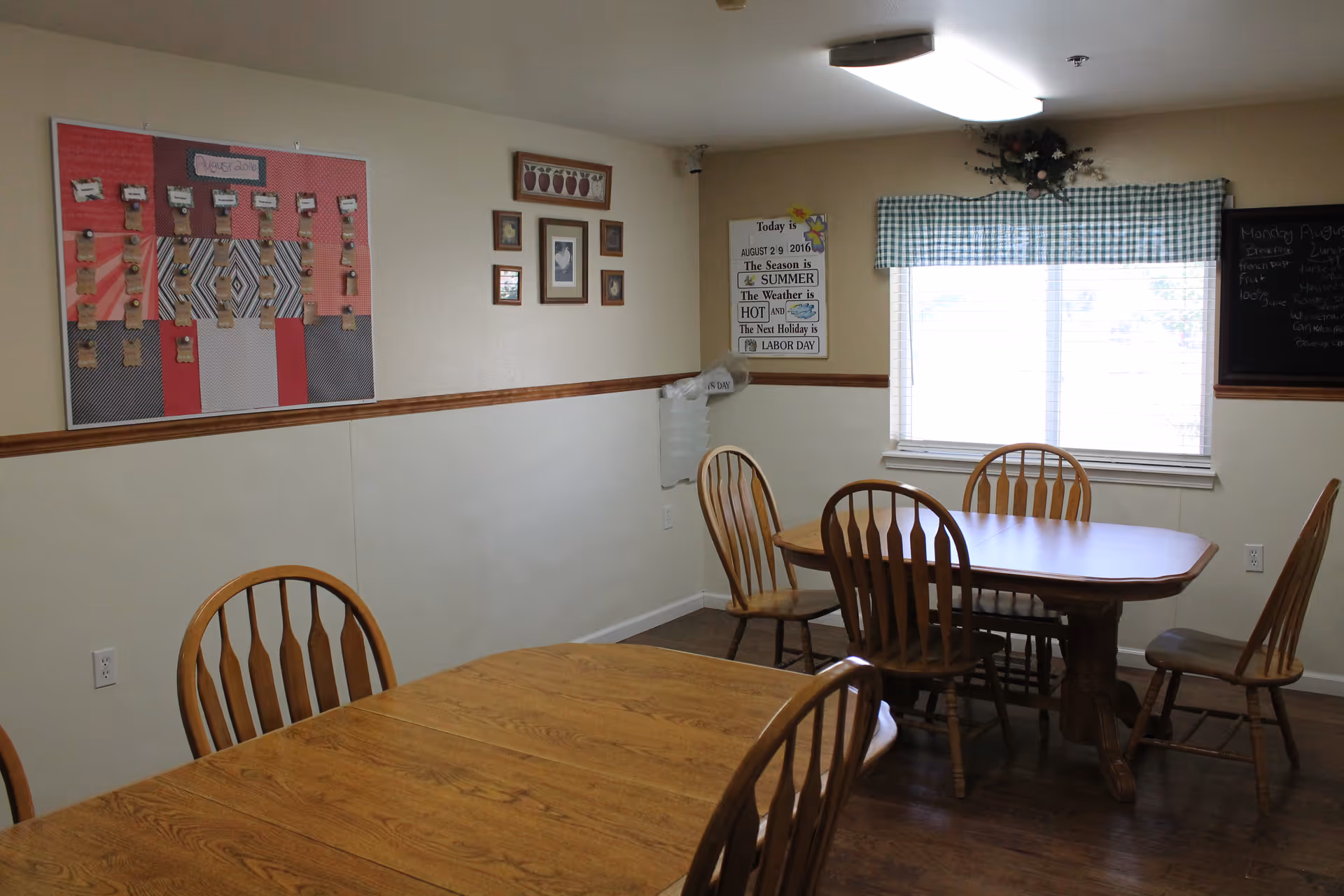 Dining room with wooden tables and chairs, a bulletin board on the wall, and a window with a green gingham valance.