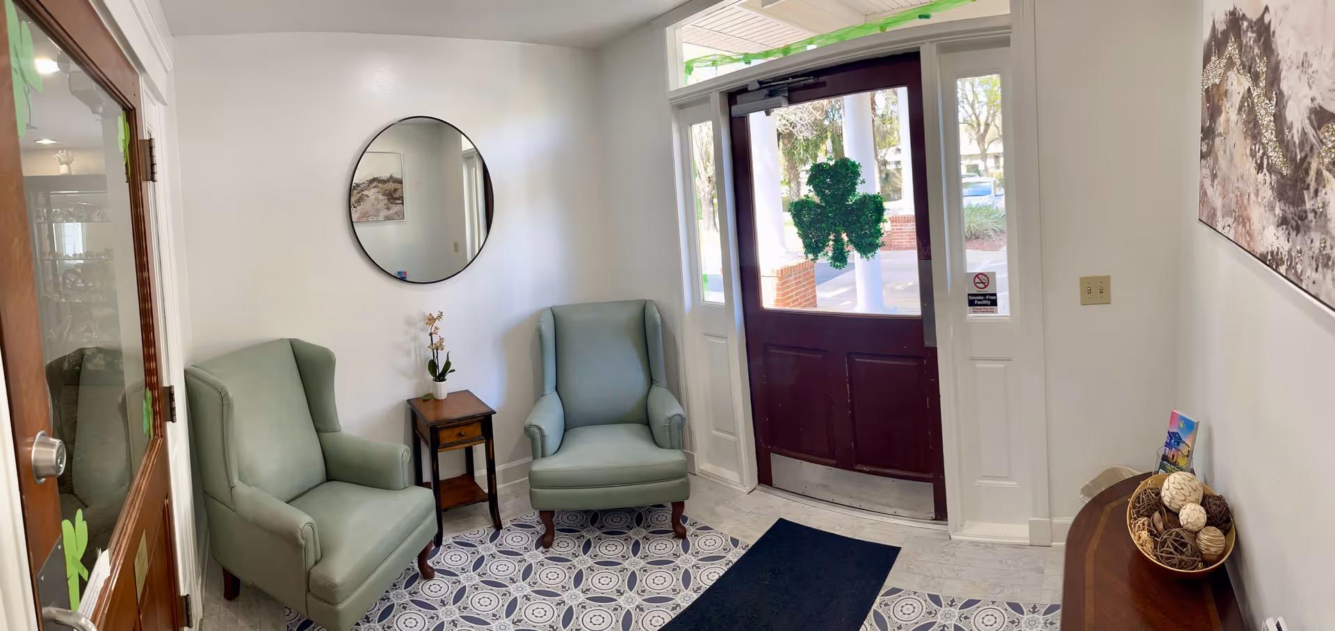 Small waiting area with two light green armchairs separated by a wooden side table with a small potted plant. A round mirror hangs on the white wall above the table. To the right is a dark wooden door with glass panels and a shamrock decoration hanging on it. A patterned rug covers part of the light-colored floor. On the right side, there is a wooden console table with decorative items and a colorful card. The space is well-lit with natural light coming through the door's glass panels.