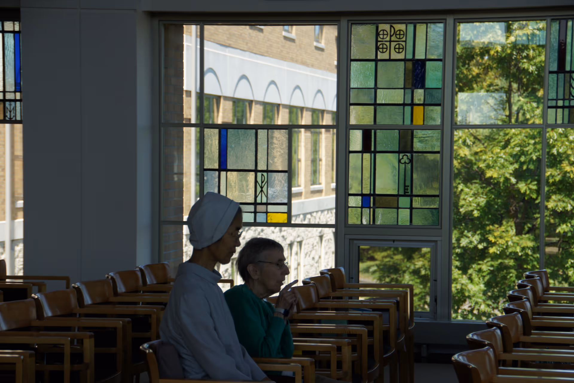 Two elderly women sitting in a room with wooden chairs arranged in rows. One woman is wearing a white head covering and the other has short gray hair and glasses. The room has large windows with decorative stained glass panels and a view of trees and a building outside.