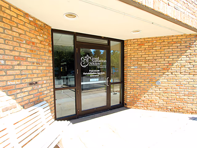 Glass door entrance to Good Samaritan Society - Stillwater Post-Acute Rehabilitation Services, set in a brick building exterior with a wooden bench nearby.
