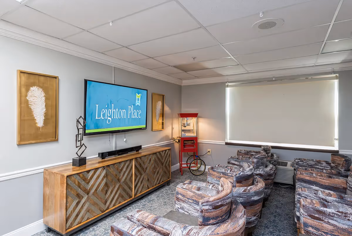 A small theater-style room with multiple patterned armchairs arranged in rows facing a wall-mounted TV screen displaying 'Leighton Place'. The room has gray walls with two framed feather artworks, a wooden cabinet below the TV, a red popcorn machine in the corner, and a window with a closed white roller shade.