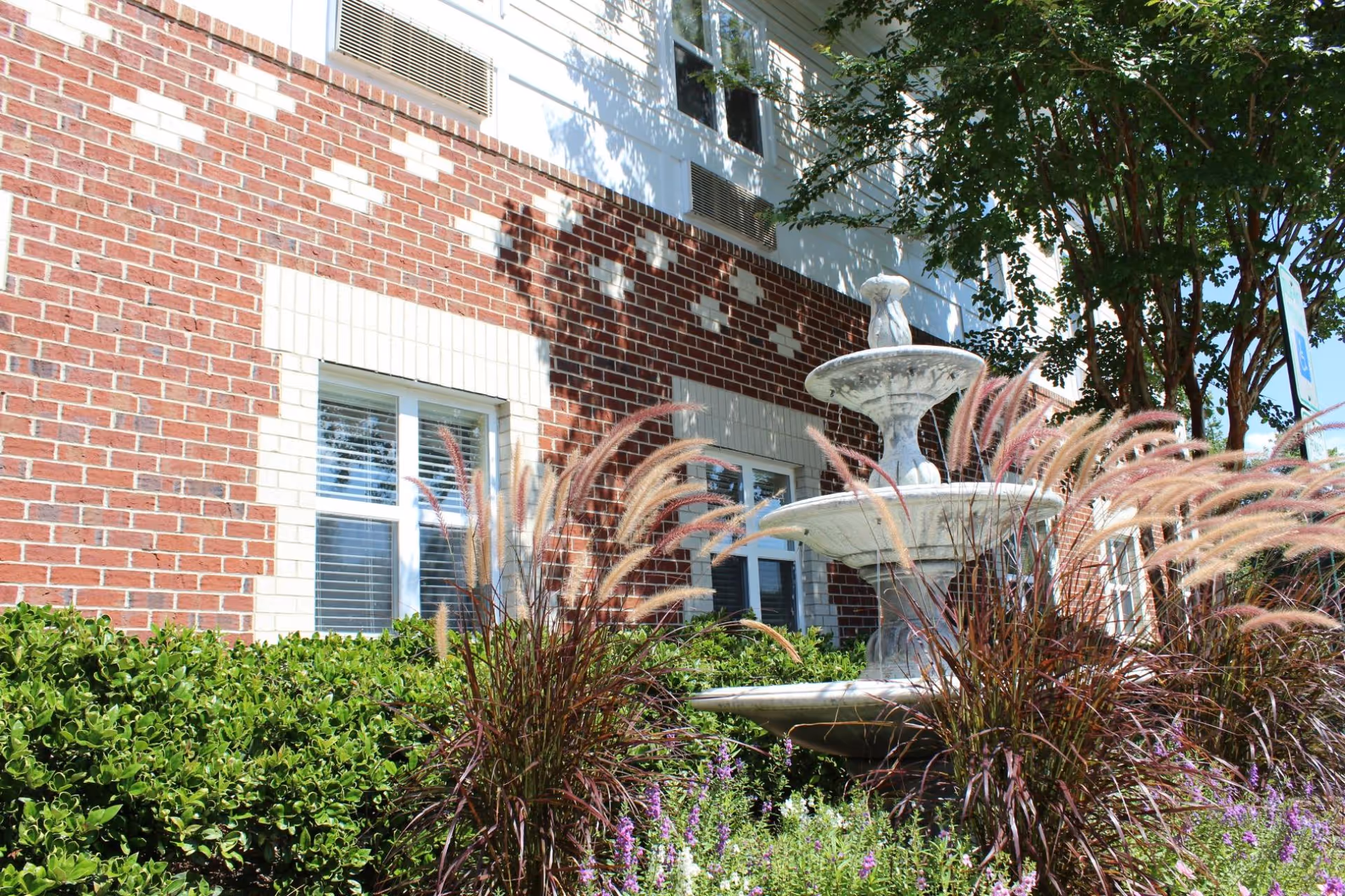 Outdoor view of a brick building with white window frames and air conditioning units. In front of the building, there is a decorative stone fountain surrounded by ornamental grasses, green bushes, and flowering plants. A tree is visible on the right side providing some shade.
