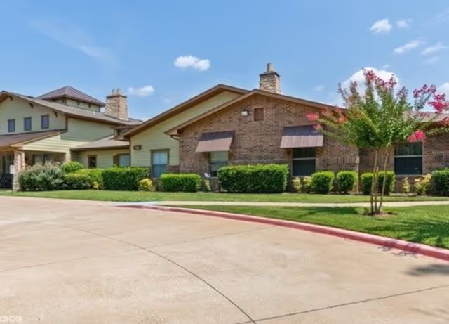 Front exterior of a single-story brick assisted living building with shrubs, a curved driveway, and a small tree under a blue sky.