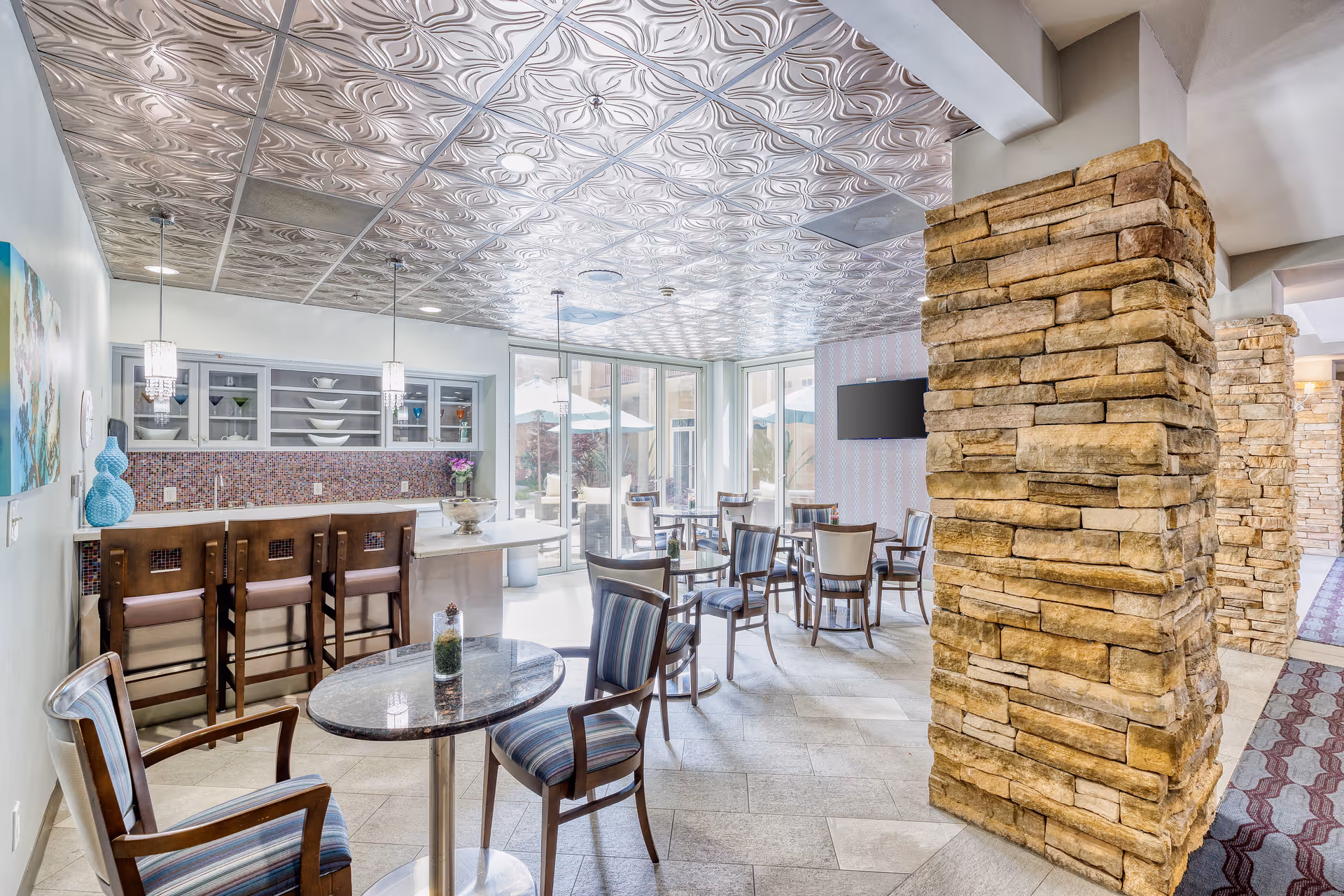 A bright and modern common area with a small bar featuring three high chairs, a round table with two chairs in the foreground, and several other tables and chairs arranged near large windows. The ceiling has an ornate metallic design, and there are stone pillars dividing the space. A flat-screen TV is mounted on the wall, and the room opens to an outdoor patio area.