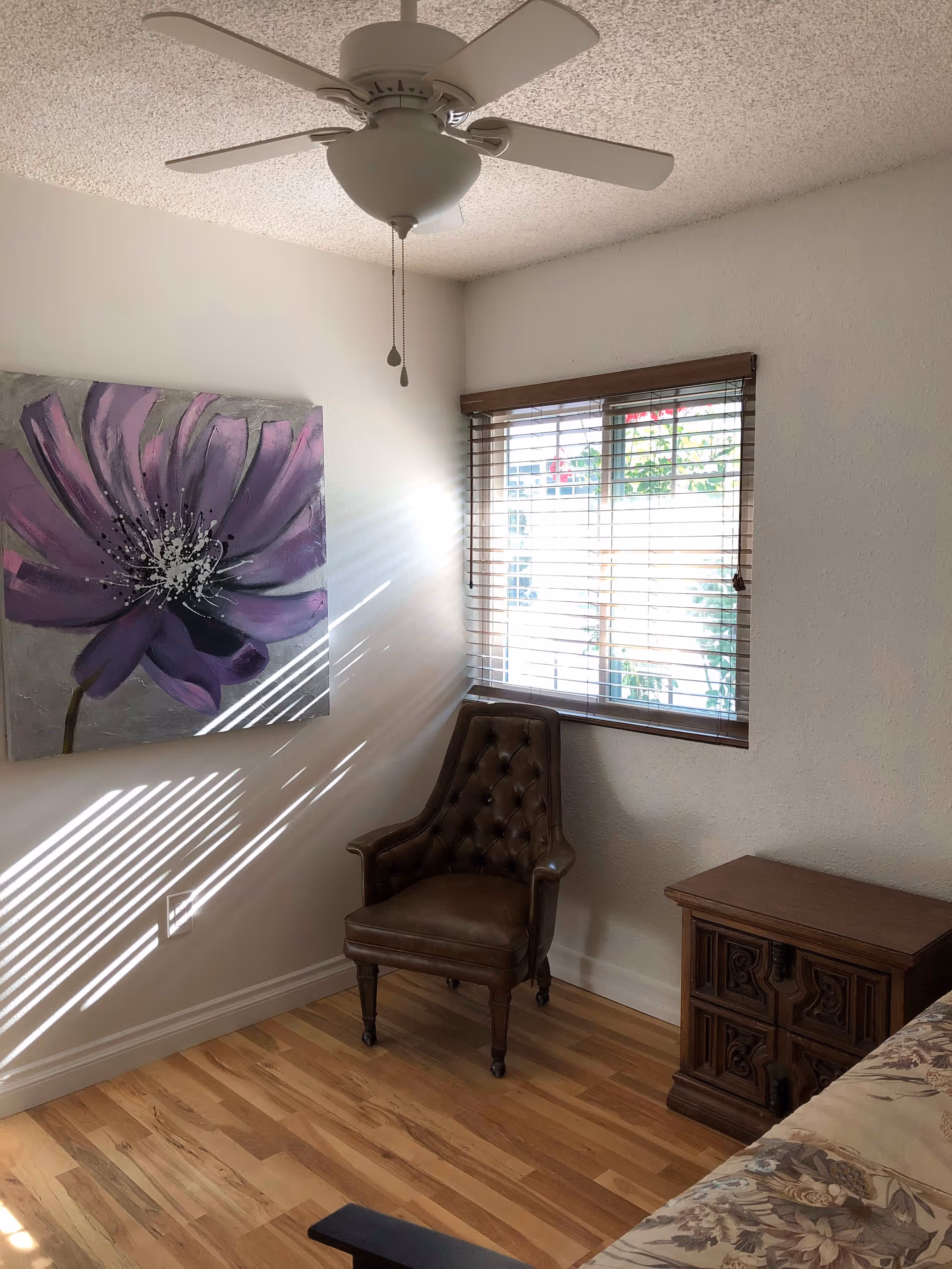 A corner of a bedroom with a brown leather tufted armchair, a wooden nightstand with carved details, a window with wooden blinds letting in sunlight, a ceiling fan with light, and a large painting of a purple flower on the wall.