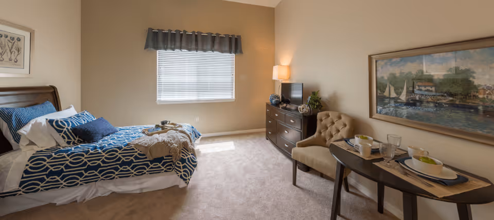 A cozy bedroom with a bed dressed in blue and white patterned bedding, a window with blinds and a valance, a dresser with a lamp and decorative items, a cushioned chair, and a small table set with dishes and cups. A large framed painting of a waterfront scene hangs on the wall.