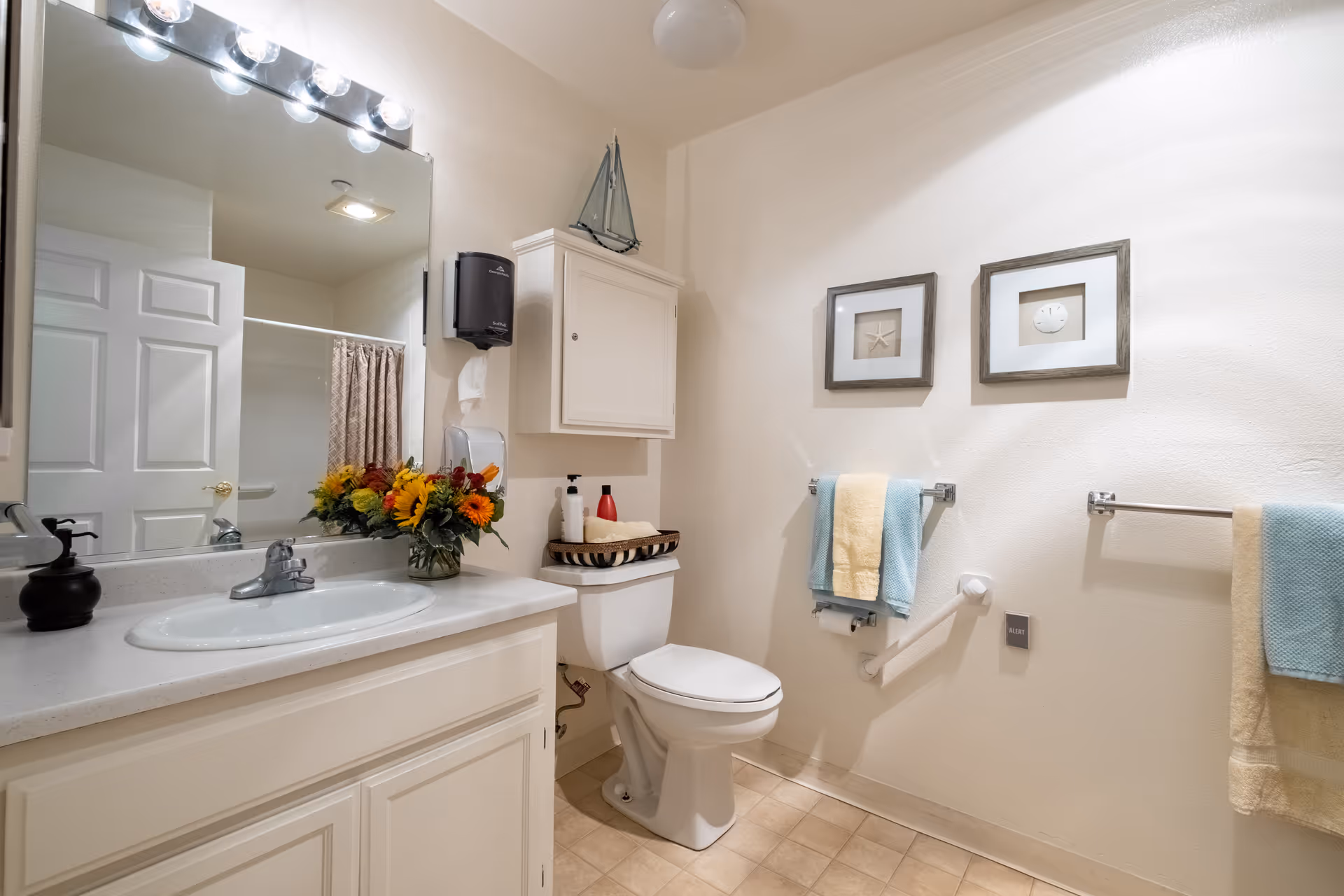 A clean and well-lit bathroom featuring a white sink with a countertop, a large mirror with lights above it, a toilet with a basket of toiletries on top, a white cabinet mounted on the wall, and two framed pictures of sea-themed items on the wall. There are also blue and yellow towels hanging on towel racks and a grab bar near the toilet.