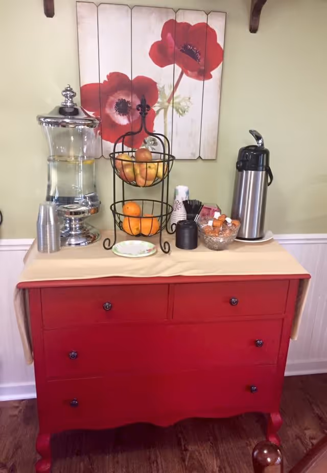 A red wooden dresser with a beige cloth on top, holding a glass water dispenser with lemon slices, a two-tiered fruit basket with apples and oranges, a stainless steel coffee dispenser, disposable cups, stirrers, and a bowl of sugar packets. Behind the dresser is a wall with a painting of two large red flowers on white wooden panels.