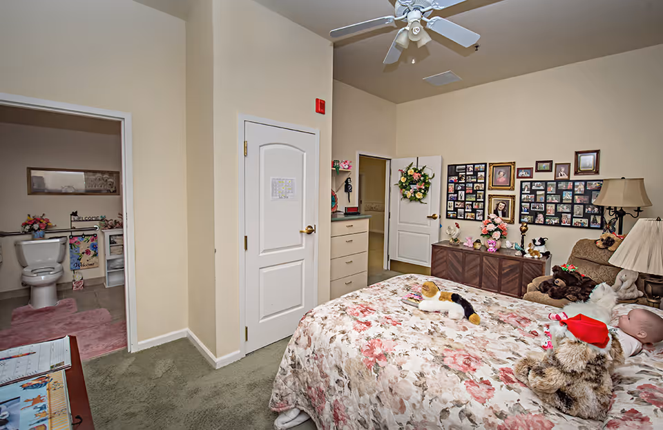 A decorated bedroom in a senior living facility with a floral bedspread, stuffed animals, a photo collage on the wall, and an open door showing an en-suite bathroom.