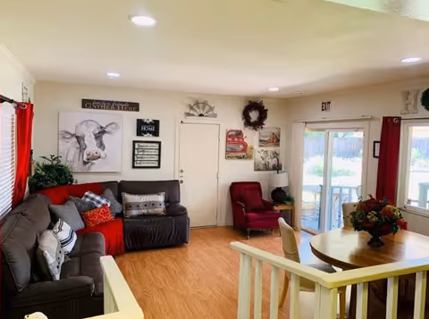 A cozy living room area with a dark gray sectional sofa adorned with various pillows and a red throw blanket. There is a red armchair near a white door, and several decorative wall hangings including a cow painting, wreath, and framed quotes. A round wooden table with a floral centerpiece is positioned near sliding glass doors with red curtains, leading to an outdoor patio. The room has light-colored walls and wood flooring.