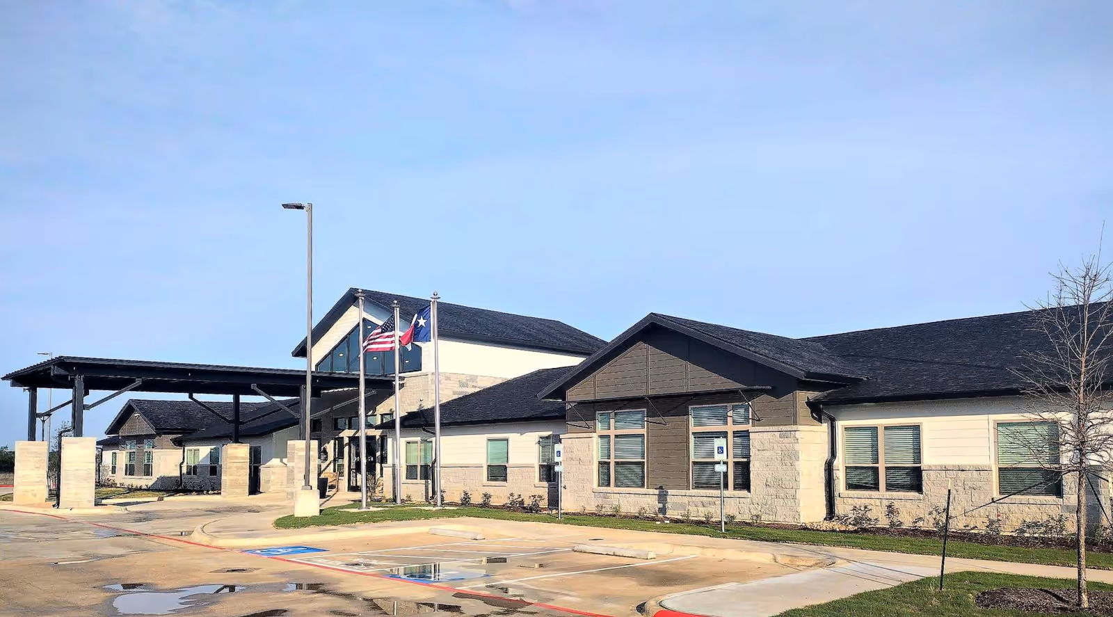 Exterior view of a single-story building with a covered entrance, three flagpoles displaying the American flag, Texas state flag, and another flag, and a parking lot with handicap spaces in front. The building has a combination of stone and siding exterior with multiple windows and a clear blue sky above.