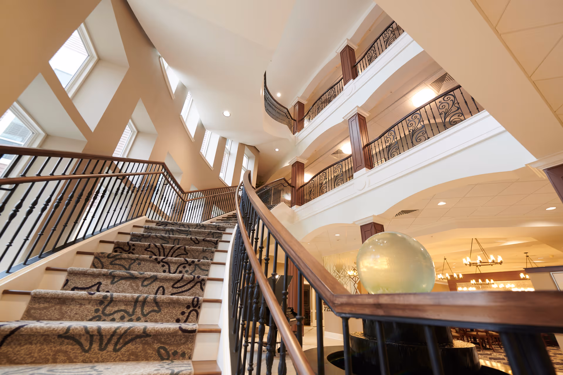 Interior view of a senior living facility showing a carpeted staircase with ornate black railings leading up to multiple floors with balconies. The area is well-lit with natural light from tall windows and ceiling lights. A decorative glass orb is placed on the railing in the foreground.
