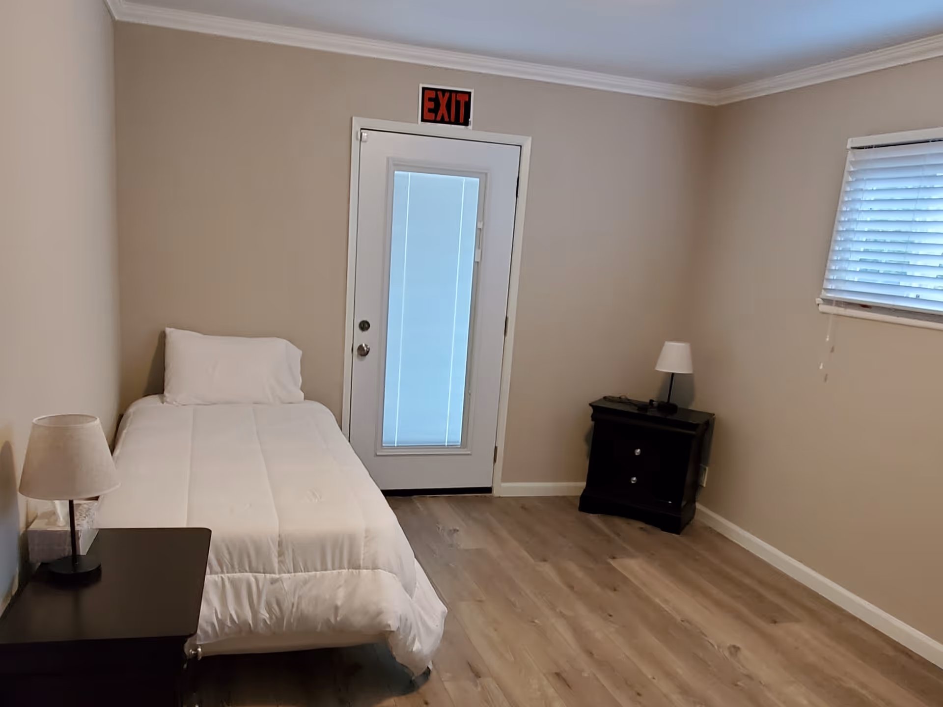 A small bedroom with a single bed covered in white bedding, a nightstand with a lamp on each side of the bed, a window with blinds on the right wall, and a door with a glass panel and an exit sign above it.