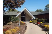 Entrance walkway leading to the front door of Westridge Health Care Center, surrounded by landscaped bushes, flowers, and trees under a clear blue sky.