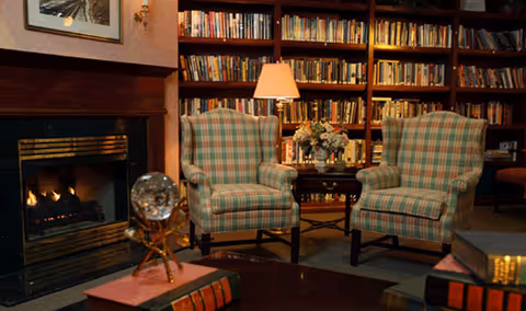 Cozy library-style living room with two plaid armchairs facing a small table, a lit fireplace and bookshelves in the background.