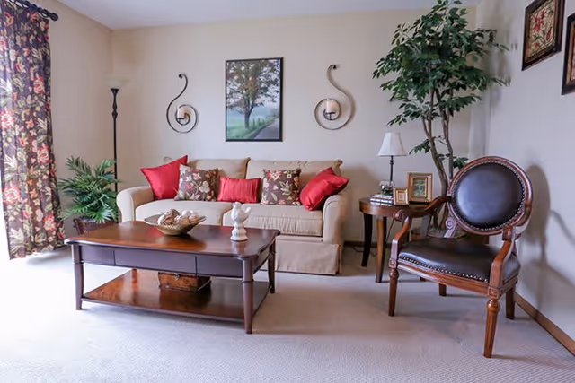 A cozy living room with a beige sofa adorned with red and floral cushions, a wooden coffee table with decorative items, a wooden armchair with dark leather upholstery, a side table with a lamp and picture frames, a tall floor lamp, a potted plant, and framed artwork on the walls.