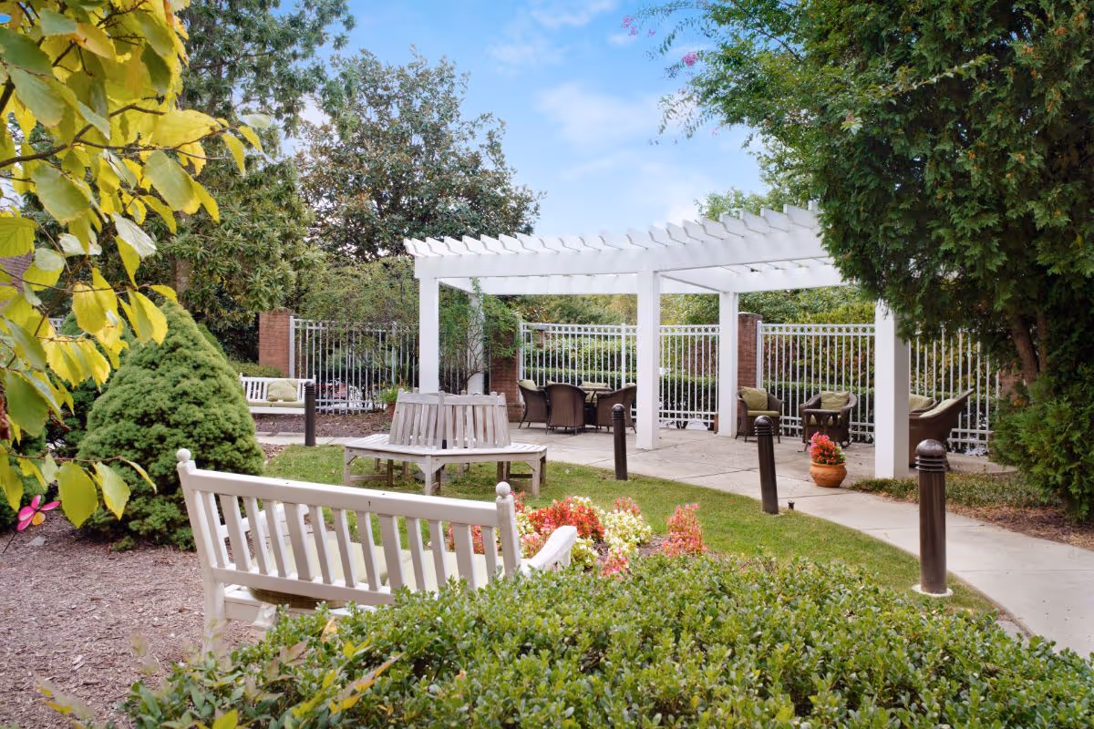 Outdoor courtyard with a white pergola, benches, chairs and landscaped shrubs and flowers.