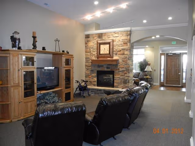 A cozy living room area in an assisted living facility featuring a stone fireplace with a clock above it, a wooden entertainment center with a TV, and several black leather recliners arranged facing the fireplace. A walker is positioned near the fireplace, and the room has a neutral color palette with soft lighting. The entrance door is visible in the background.