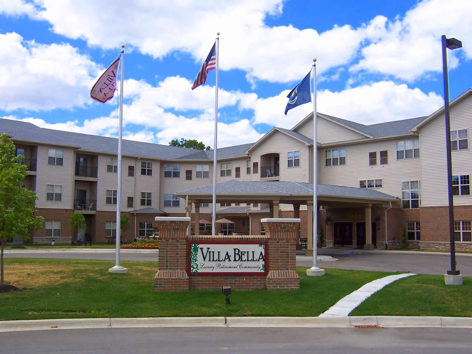Exterior view of Villa Bella, a luxury retirement community building with three stories, beige siding, and brick accents. There are three flagpoles with flags in front of the entrance, and a sign that reads 'Villa Bella Luxury Retirement Community' on a brick base. The sky is partly cloudy and the surrounding area is landscaped with grass and small trees.