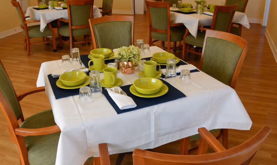 A dining room table set with green plates, bowls, mugs, and clear glasses on a white tablecloth with dark blue placemats. The table has four wooden chairs with green cushions. In the background, there are more similarly set tables and chairs on a wooden floor.