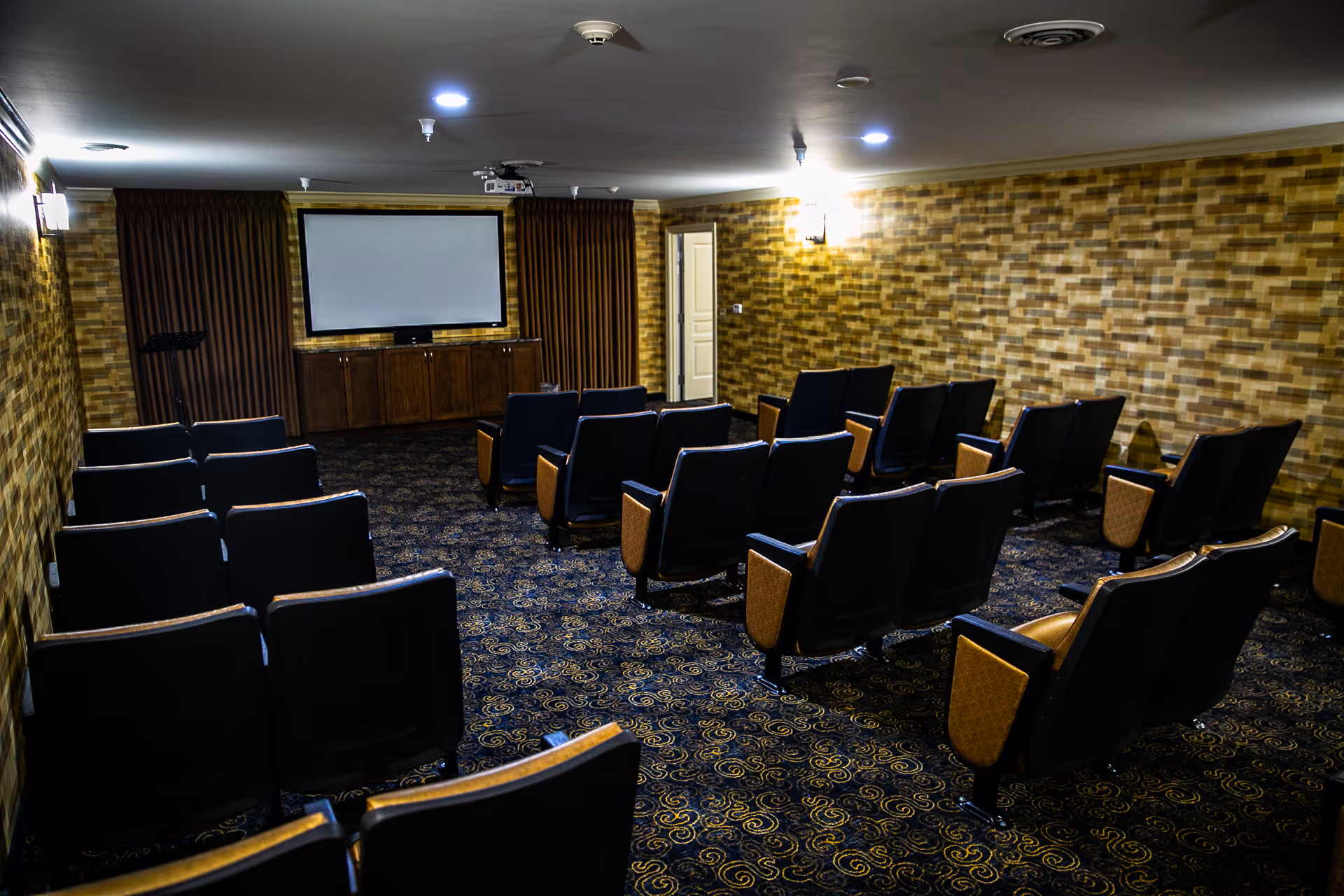 Small movie-theater style room with rows of upholstered seats facing a projection screen, patterned carpet and wood-paneled walls.