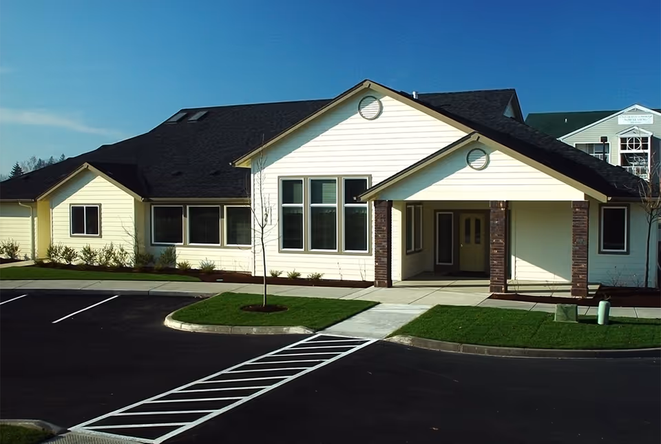 Exterior view of a single-story building with a black roof and light yellow siding, featuring a covered entrance supported by brick columns, surrounded by a parking lot and landscaped grass areas under a clear blue sky.