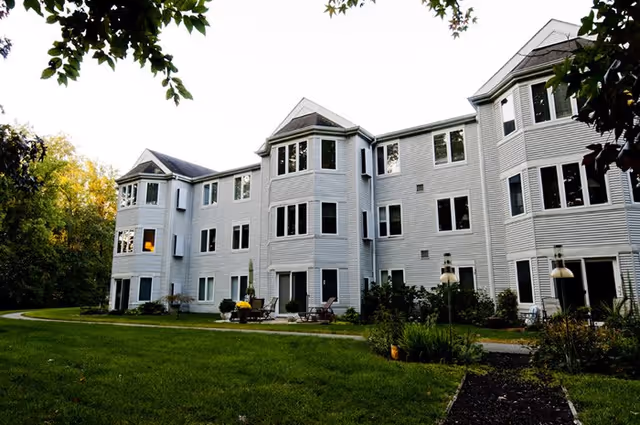 Exterior view of a three-story white senior living facility building with multiple windows and a well-maintained lawn and garden area in front, surrounded by trees.