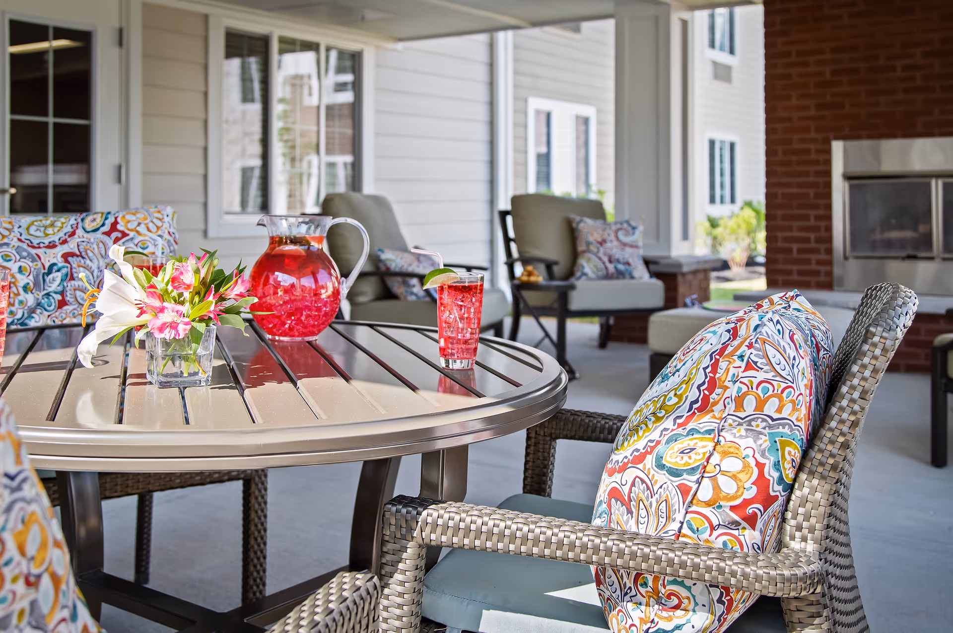 Outdoor patio area with a round table featuring a pitcher and glass of red beverage with a lime wedge, and a small vase of flowers. Surrounding the table are wicker chairs with colorful patterned cushions. In the background, cushioned armchairs and a brick fireplace are visible.