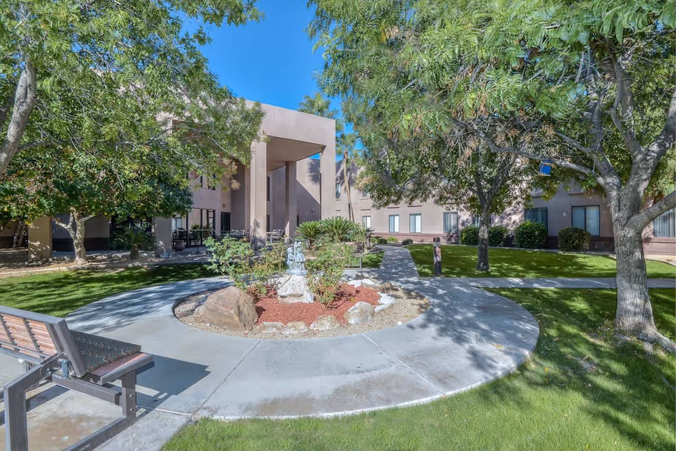 Outdoor courtyard area of Desert Winds Assisted Living featuring a circular concrete walkway surrounding a small garden with rocks and plants. There are benches along the walkway, green grass, several trees providing shade, and a beige building with large windows in the background under a clear blue sky.