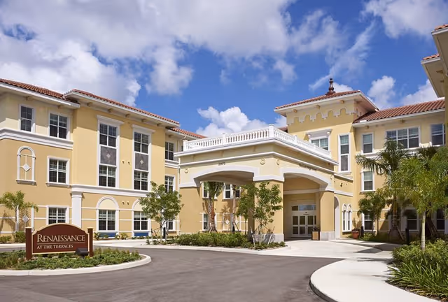 Exterior view of a large, yellow senior living facility building with white trim and a covered entrance. The building has multiple windows and a red-tiled roof. There are palm trees and landscaped greenery around the entrance and along the driveway under a partly cloudy blue sky.