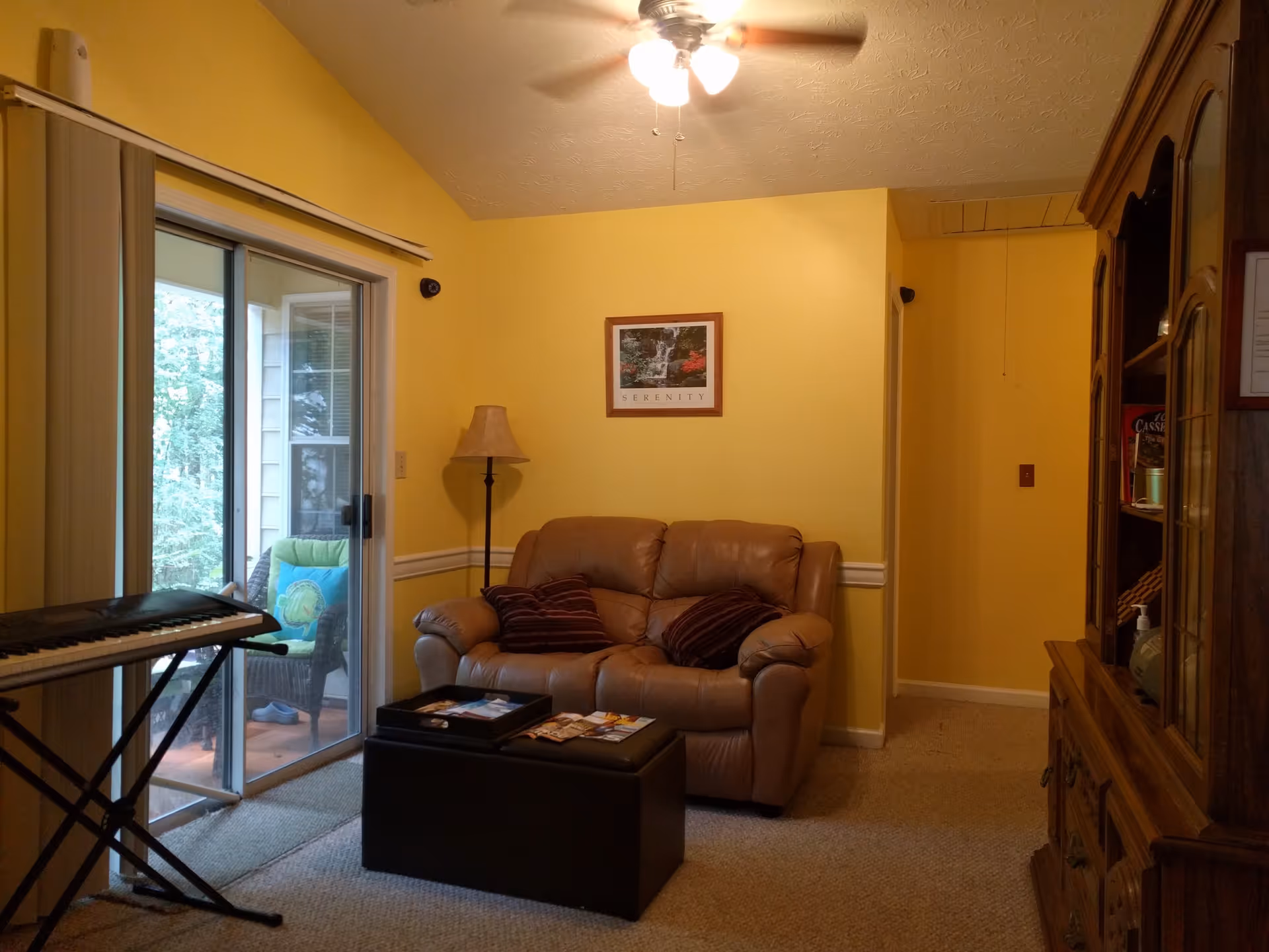 A cozy living room with yellow walls, a ceiling fan with lights, a brown leather loveseat with two striped pillows, a floor lamp, a black ottoman with magazines on top, a wooden cabinet with glass doors, and a keyboard on a stand near sliding glass doors leading to a small outdoor patio with a wicker chair and green cushions.