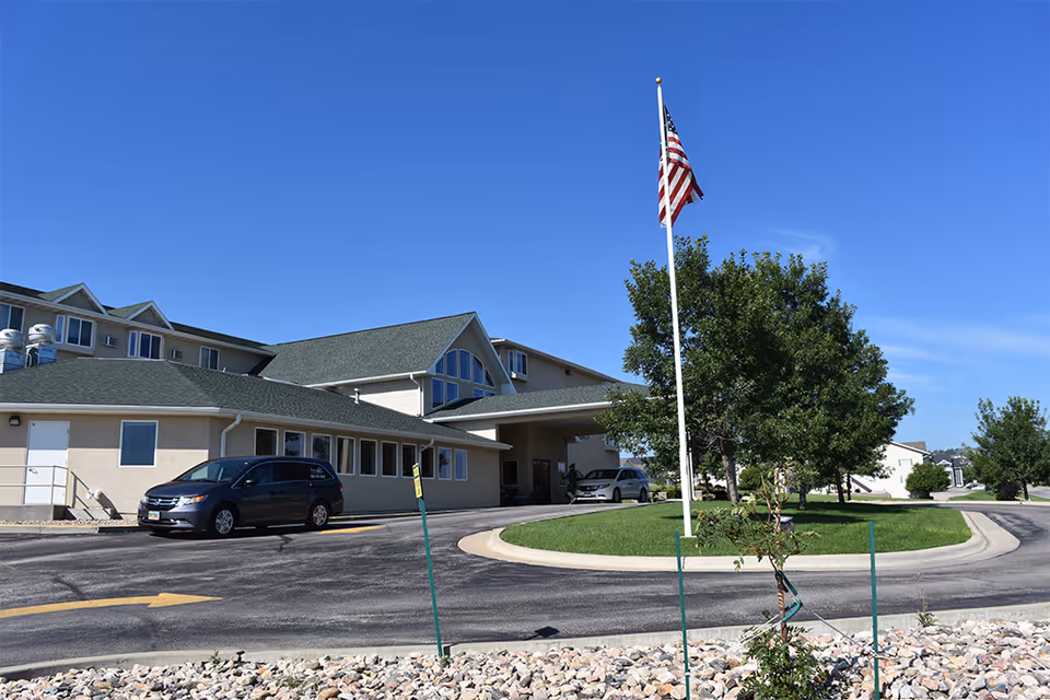 Exterior view of a senior living facility building with a green roof and beige walls under a clear blue sky. There is a driveway with parked cars, a circular grassy area with a tree and an American flag on a flagpole in the center.