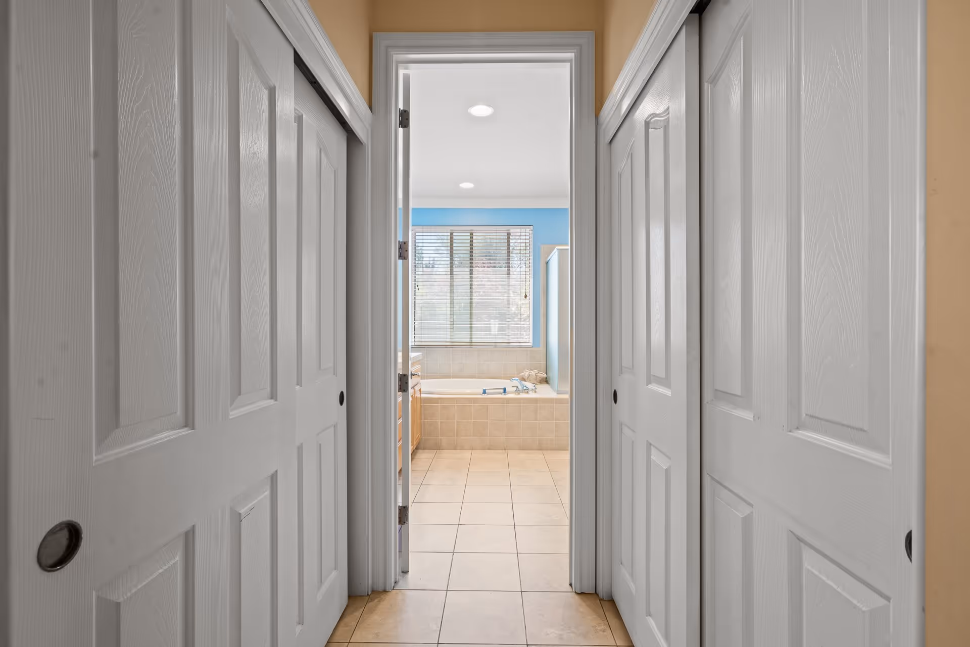 View down a hallway with white sliding closet doors on both sides leading to a bathroom with a large bathtub, tiled floor, and a window with blinds. The bathroom walls are painted blue.