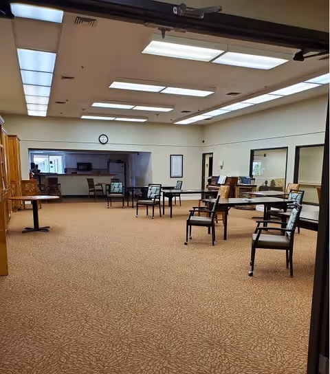 A spacious room with carpeted floor and several tables and chairs arranged sparsely. The ceiling has multiple rectangular fluorescent light panels. At the far end, there is a kitchen area with a refrigerator, microwave, and cabinets. A clock is mounted on the wall above the kitchen opening. The room has large windows on the right side and a few framed items on the walls.