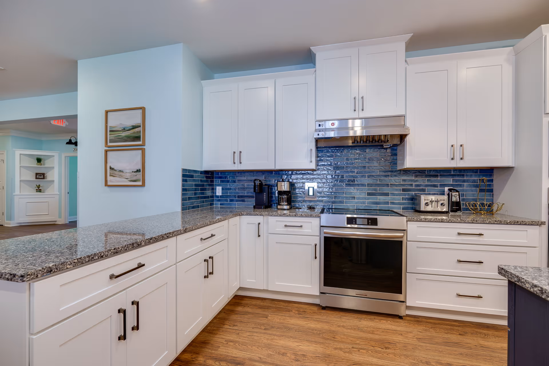 Modern kitchen with white cabinets, granite countertops, and a blue tile backsplash. Stainless steel oven and range hood are centered between the cabinets. Coffee maker, toaster, and other small appliances are on the countertops. The floor is wood, and two framed landscape paintings hang on a light blue wall.